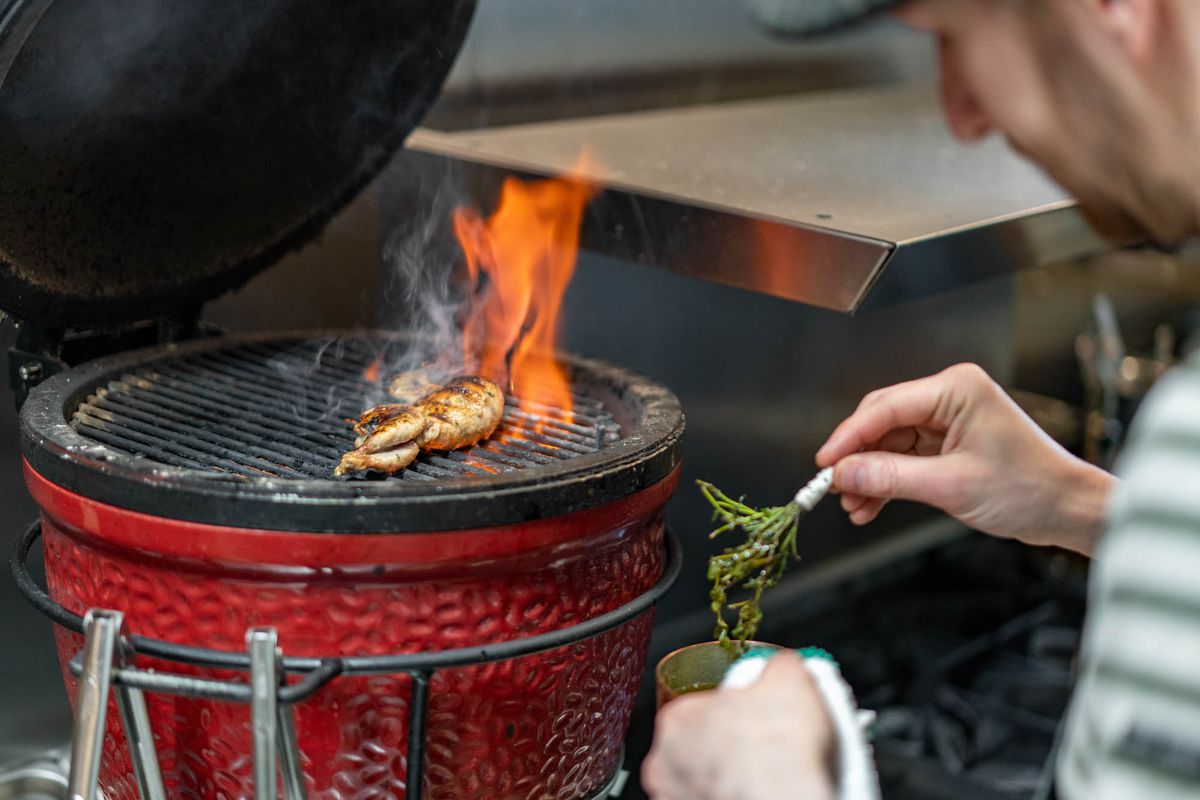A whole quail lays on a grill while a man brushes it with a thyme sprig.