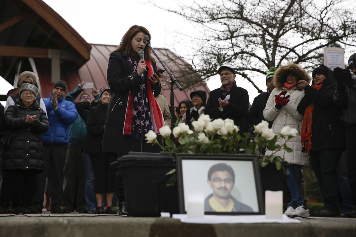 A woman speaks at a vigil behind a bouquet of white flowers and a photo of Srinivas Kuchibhotla.