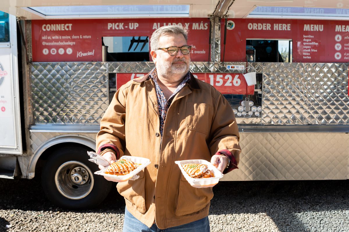 Steve Sando wears a brown jacket and holds two plates of tacos in front of a taco truck.
