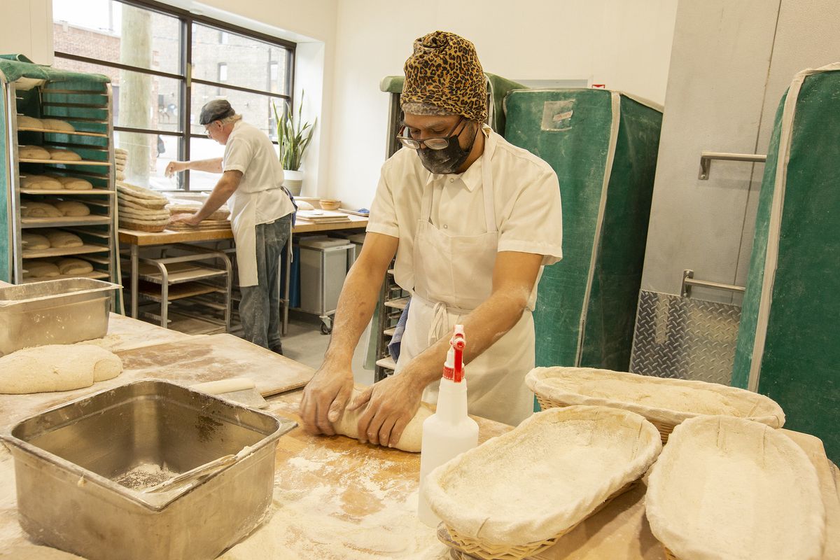 A baker stands at a table and shapes bread dough into an oblong loaf.