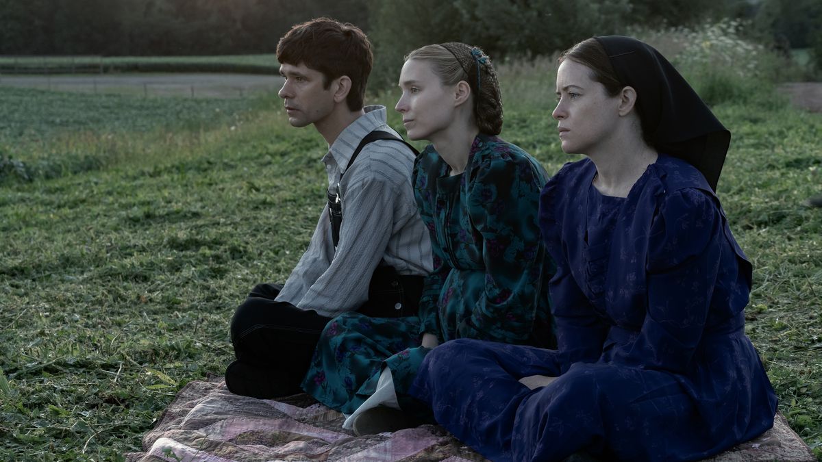 A man (Ben Whishaw), a woman (Rooney Mara) and another woman (Claire Foy) sit on a blanket and look out over a green grass field.
