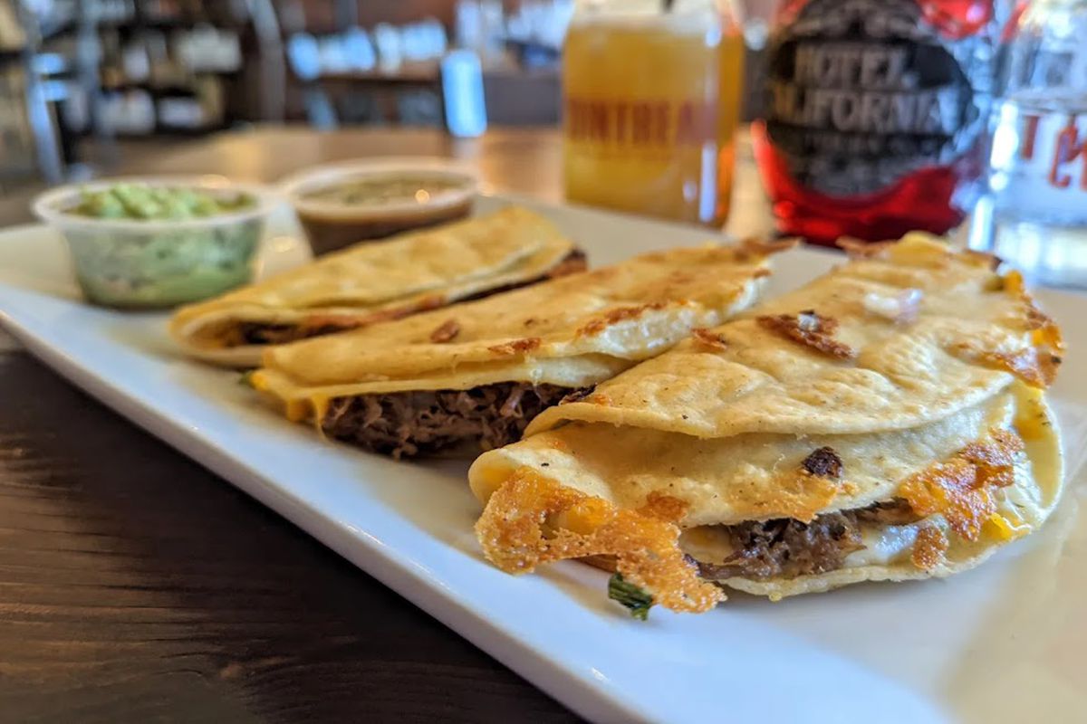 Three beef and cheese tacos in soft double shells sit on a rectangular white plate. In the background, two small plastic cups hold guacamole and a dark broth.