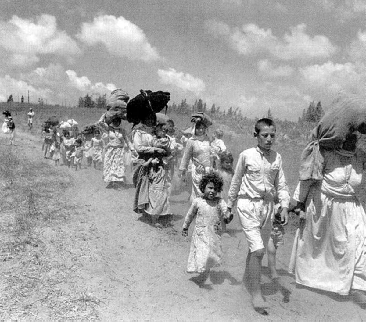 Women and children dressed in white, some carrying bundles on their heads, walk in a long line down a sandy road. 