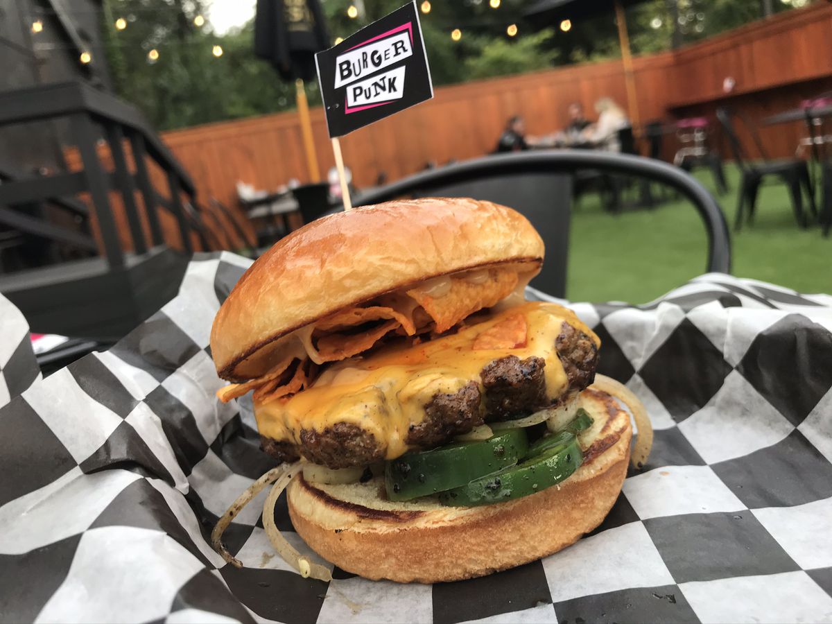 A cheeseburger topped with Doritos chips on checkerboard wax paper in front of an astroturfed outdoor seating area