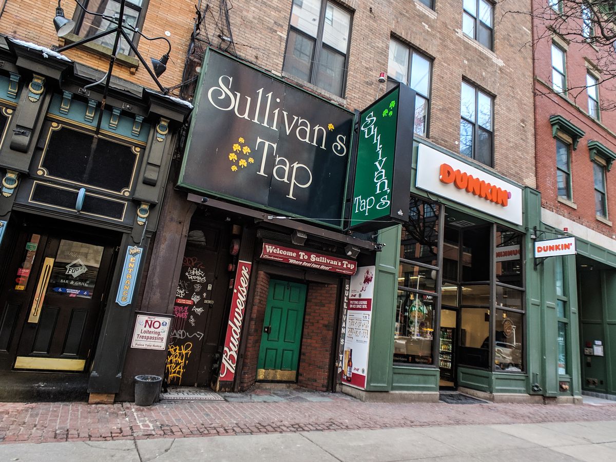 Exterior of a dive bar on a city street with a large green sign that says Sullivan’s Tap. There’s a Dunkin’ next door.