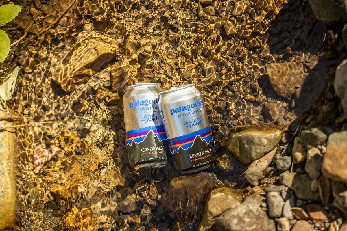 Two cans of Patagonia Provisions Dogfish Head Kernza Pils sit against some rocks in a shallow pool of water.