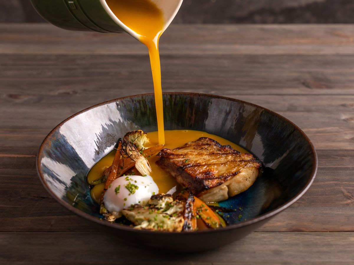 An unseen server pours broth from a serving pitcher into a bowl of tambaqui fish fillet, cauliflower, carrots, and a poached egg. The glazed bowl sits on a dark wooden table.