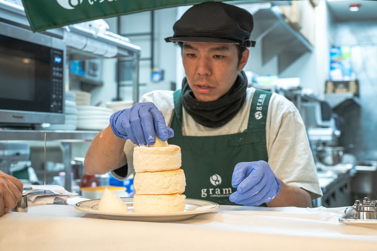 A server handles soufflé pancakes carefully, with one hand about to hold a plate and the other holding the top of a three-pancake stack.