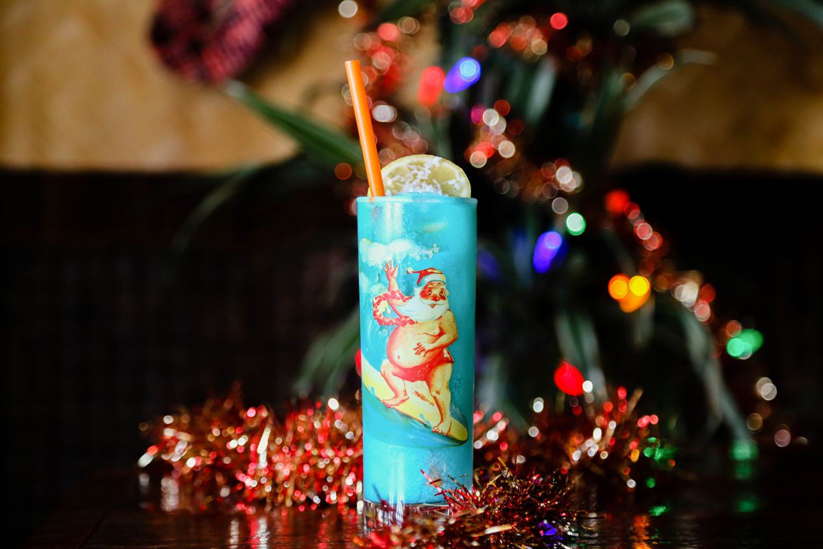 A blue cocktail in a tall glass with a surfing santa on it surrounded by tinsel.