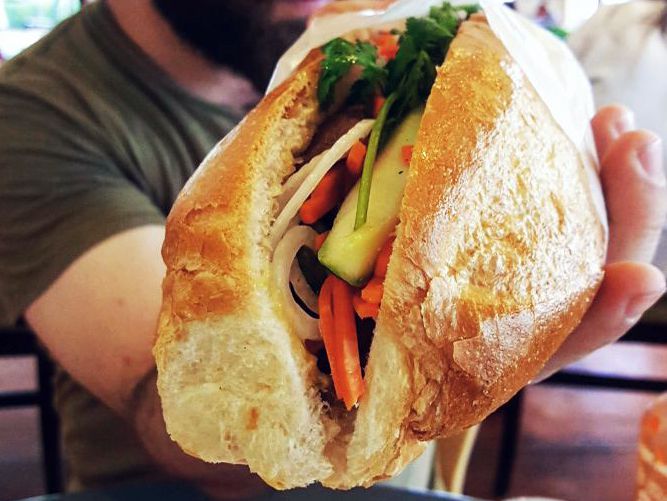 A hand holds a banh mi — crusty French bread full of vegetables —&nbsp;toward the camera
