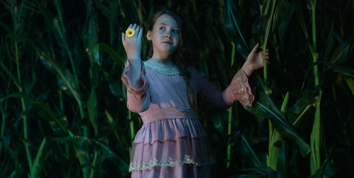 Pre-teen villain and cult leader Eden (Kate Moyer), a girl in a grubby pink dress with lace at the neck and cuffs, stands outdoors in a cornfield at night and holds up a hand sporting a bright plastic sunflower-shaped ring in the 2023 Children of the Corn