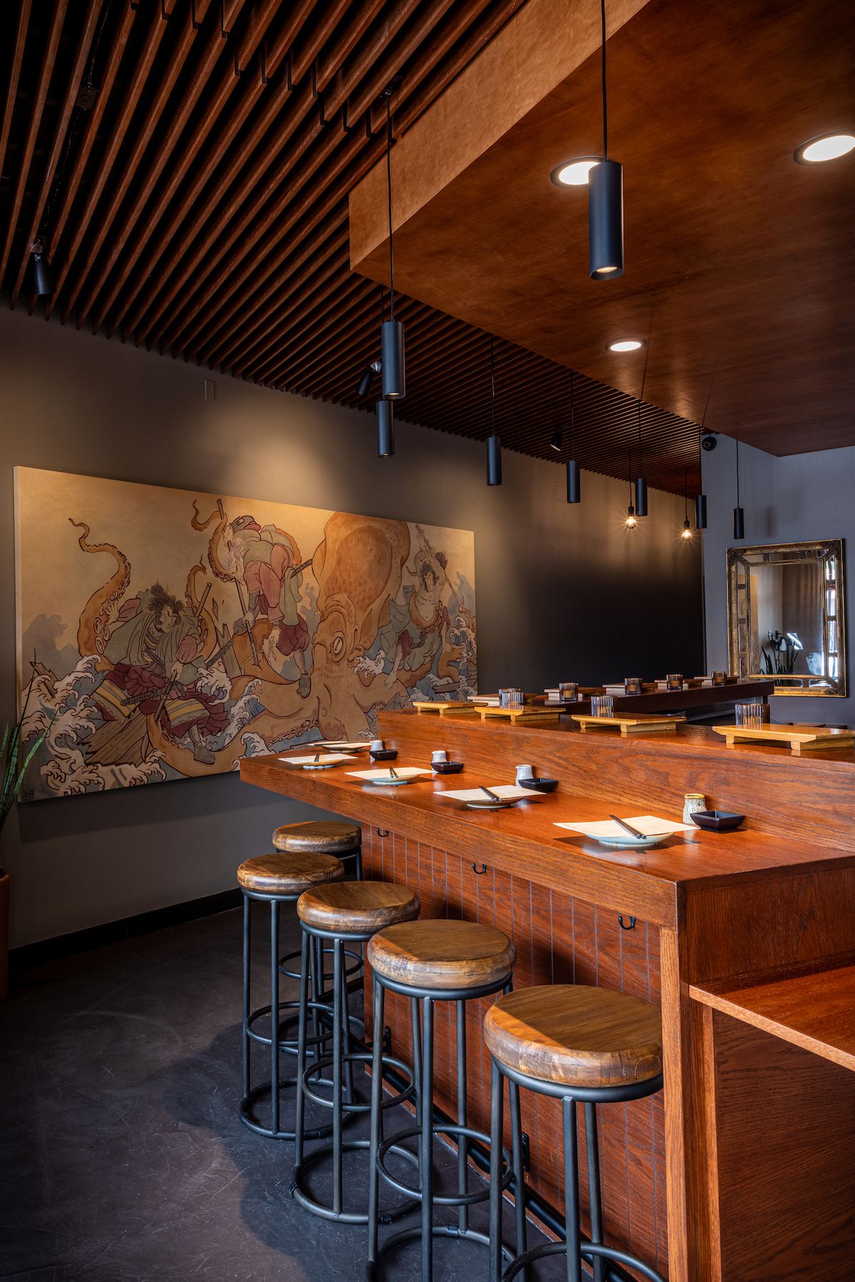 A view from one side of a tan room of a dark wooden bar with stools and art beyond at new LA sushi restaurant Santo.