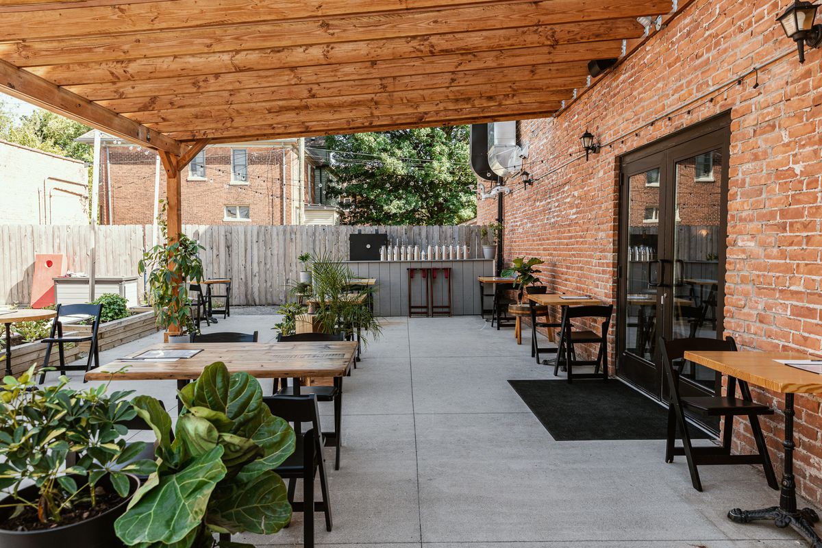 A wooden pergola covers wooden tables with folding wood chairs and plants.