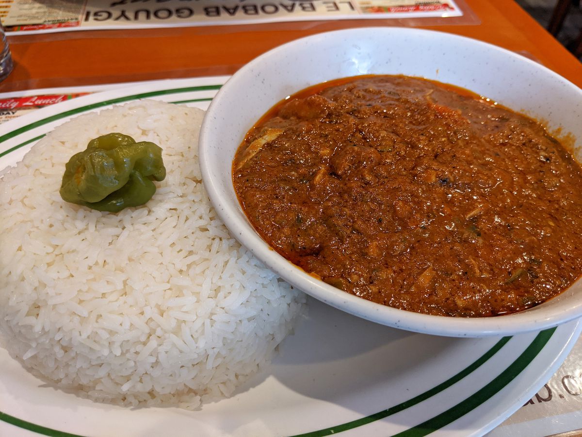 A white place with a mound of white rice on one side and a bowl with a brown chili-like dish.