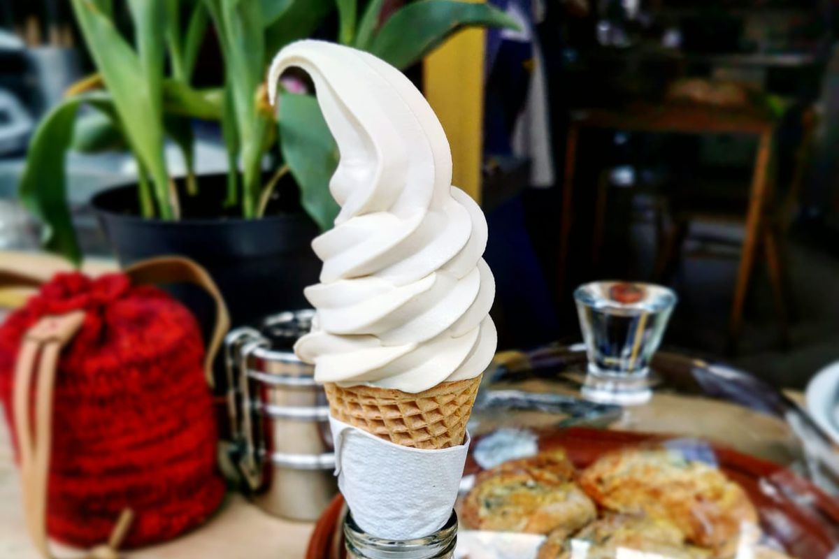 A cone of hazelnut soft serve sits in a glass bottle filled with sand, with pastries, a pot plant, and a red rattan fabric in the background