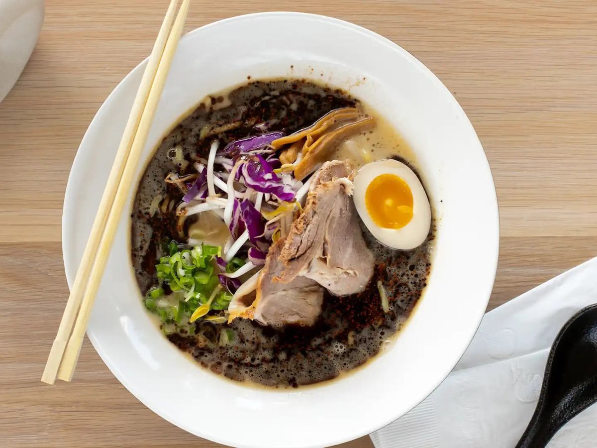 A bowl of ramen with dark broth, half hard boiled egg, meat slices, greens and veggies. 