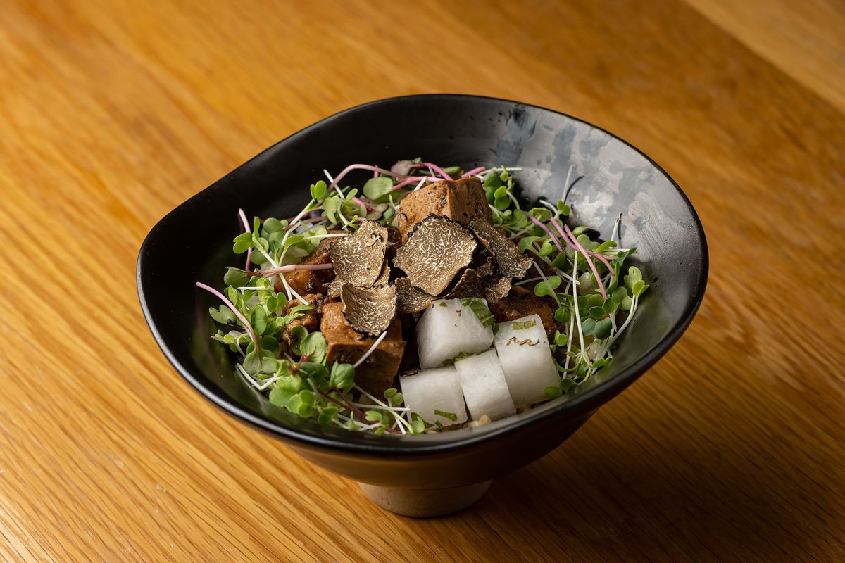 A black bowl with green leaves, cubes of white daikon, and black truffle shavings.