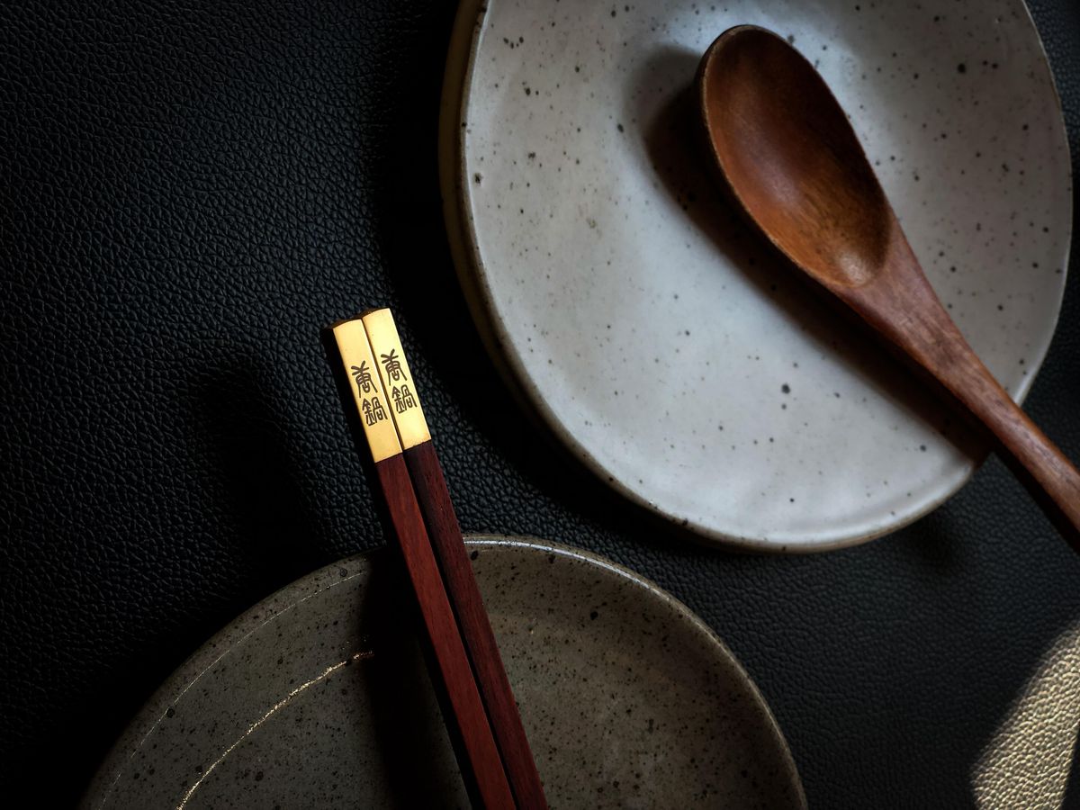 A pair of wooden chopsticks with golden ends and Chinese inscription rest on a gray plate, photographed next to a white plate and a wooden soup spoon