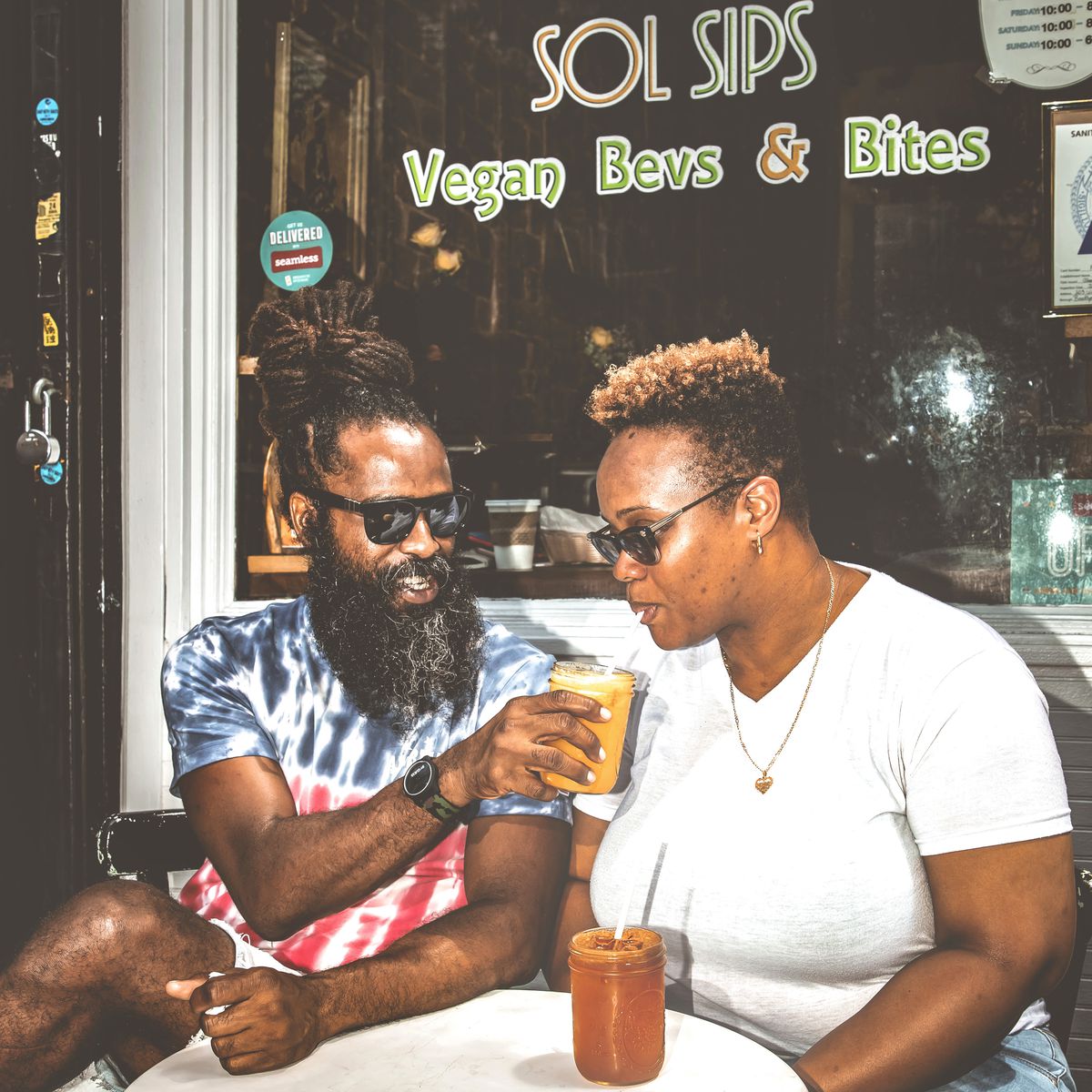 A man wearing sun glasses, with a long beard and dreadlocks, offers a woman a sip of juice in a glass jar. On the right, a woman wearing sunglasses and a white tee with short hair takes a sip.