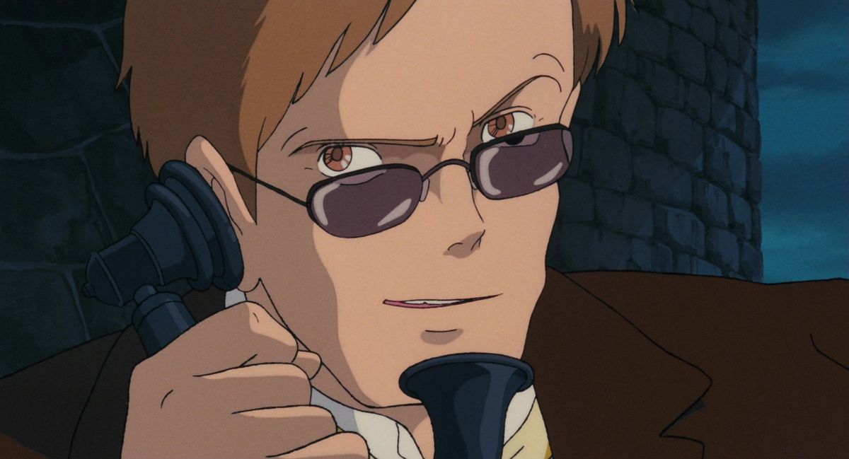 A man in brown, with brown eyes, brown hair, and brown glasses, grins as he speaks into an old-timey telephone handset in Castle in the Sky.