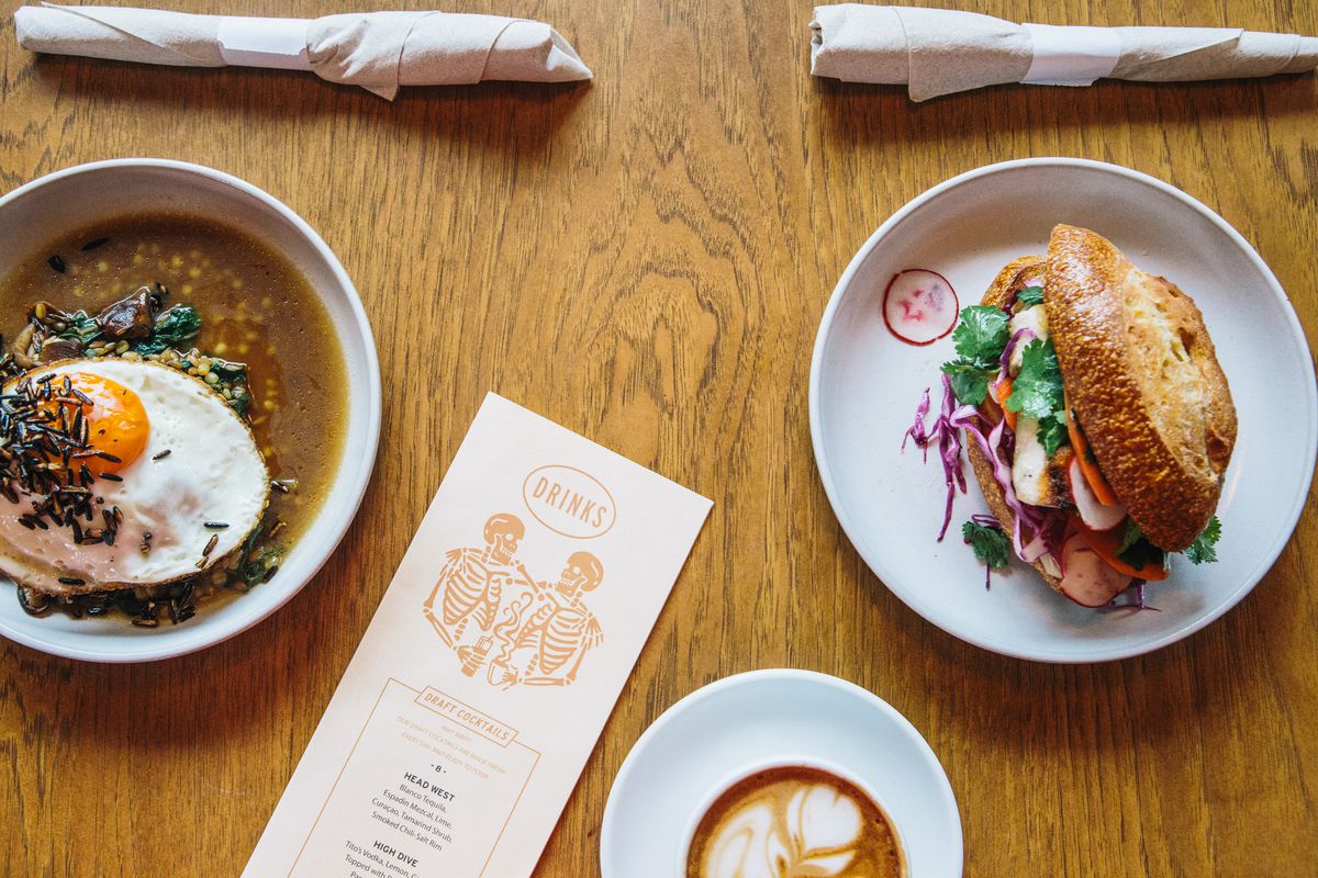 Two plates of food, one with a sunny-side-up egg and a broth and the other with a sandwich with a cup of latte and a drinks menu on a wooden table.