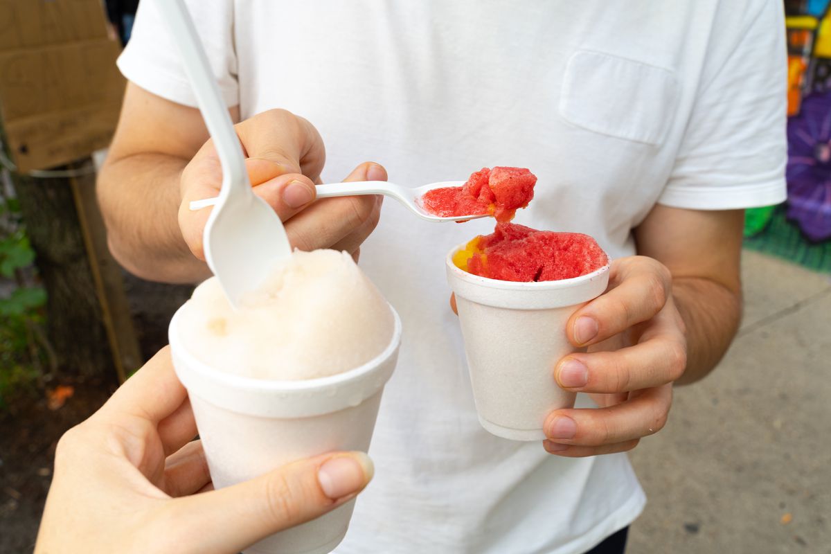 A person in a white shirt digs red Italian ice out of a styrofoam up with a plastic spoon.