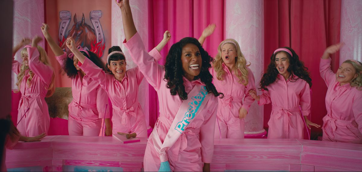 Rae, dressed in a pink jumpsuit and wearing a sash that says president, is raising one arm and appears mid-speech, while other women dressed in pink jumpsuits cheer from behind her in an all-pink room