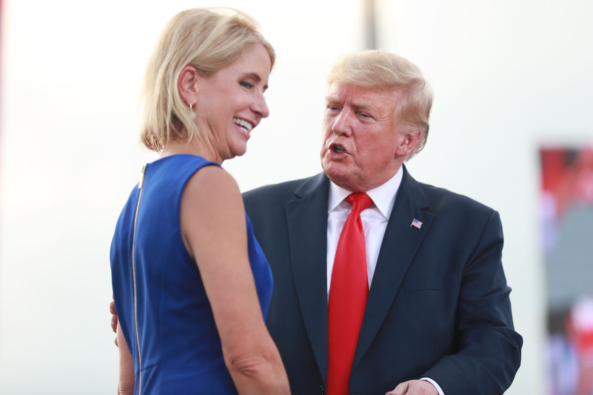 Trump, in a navy suit, white shirt, and bright red tie, is turned toward Miller, in a blue dress. They appear to be on a stage under a silver sky.