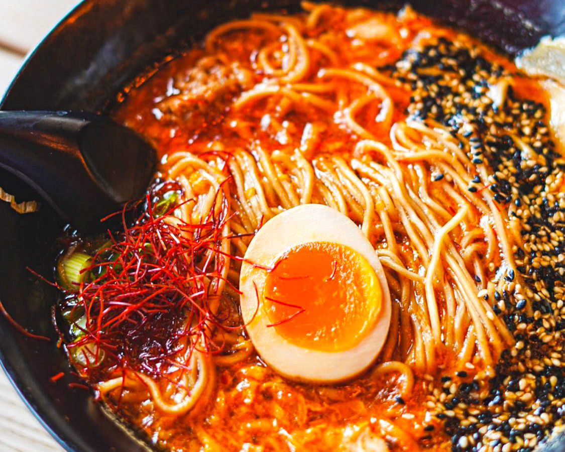 A closeup on a bowl of ramen, with squiggly noodles in a deep red broth, with egg, sesame seeds, and other fixings.