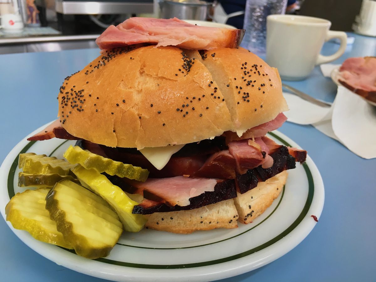 A large ham sandwich on a bun with poppy seeds. It has a slice of ham on top of the bune and a pile of pickles on the side