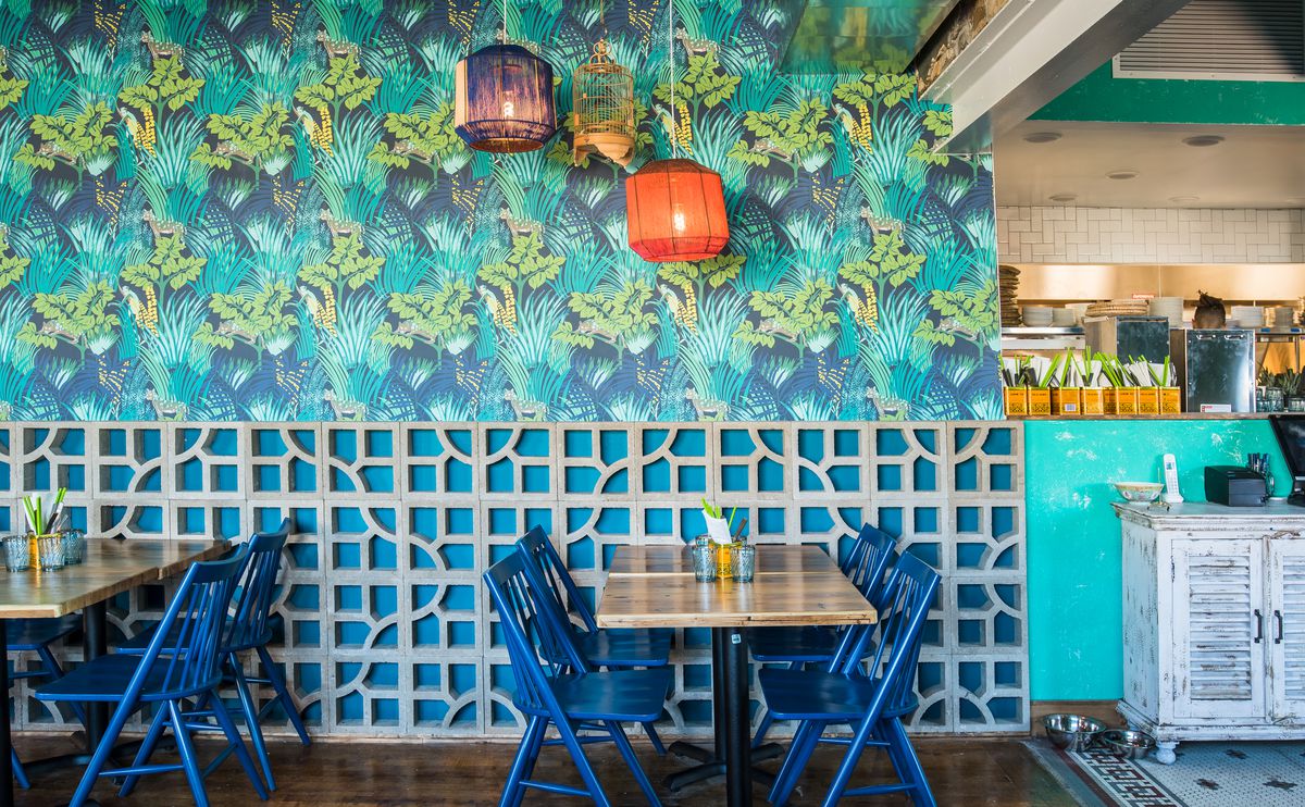 The turquoise and green wallpapered dining room at Hai Hai. Deep blue chairs are stationed at wooden tables and two orange and purple paper lanterns hang above them. 