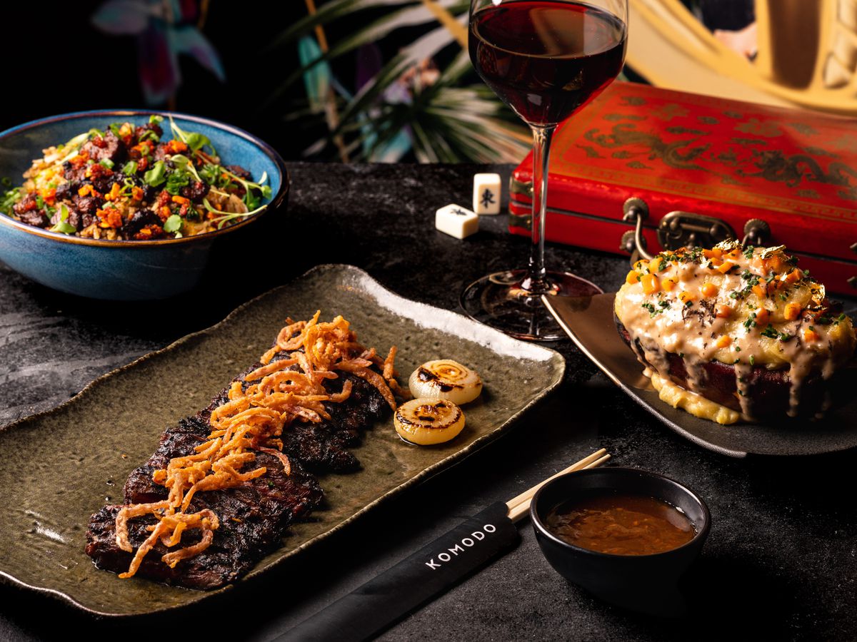 A table with lots of decor holds a silver tray with a flat iron steak covered in onions, to its right is a plate with a smothered steak, and above and to the left is a bowl of salad.