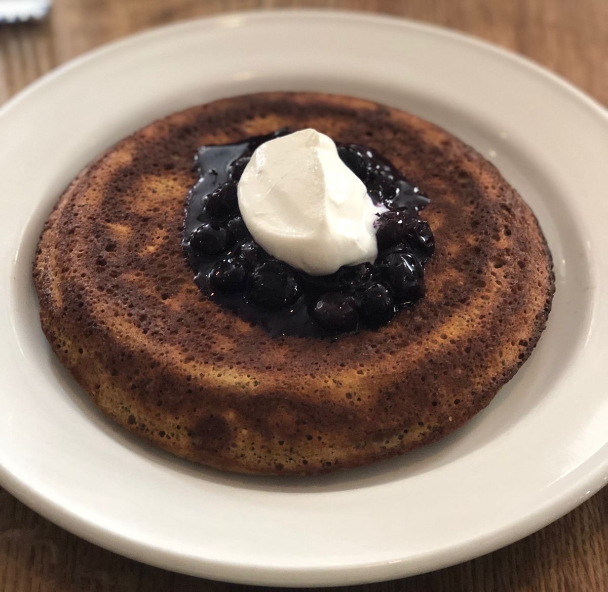 A white plate holds circular, brownish, fat pancakes with a dollop of white sour cream and purple blueberries.