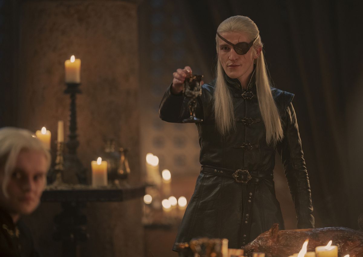 Ewan Mitchell as Aemond Targaryen in House of the Dragon raising a glass in mocking toast with his eyepatch on