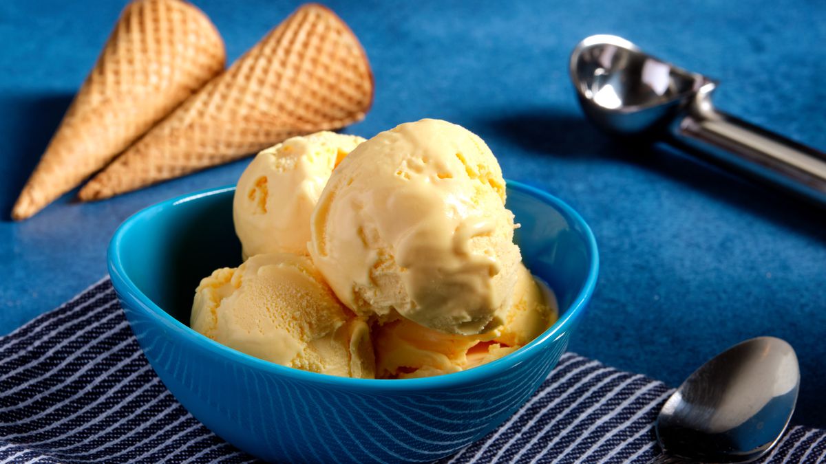 Four scoops of of mac and cheese flavored ice cream in a blue bowl, next to an ice cream scoop and two cones.