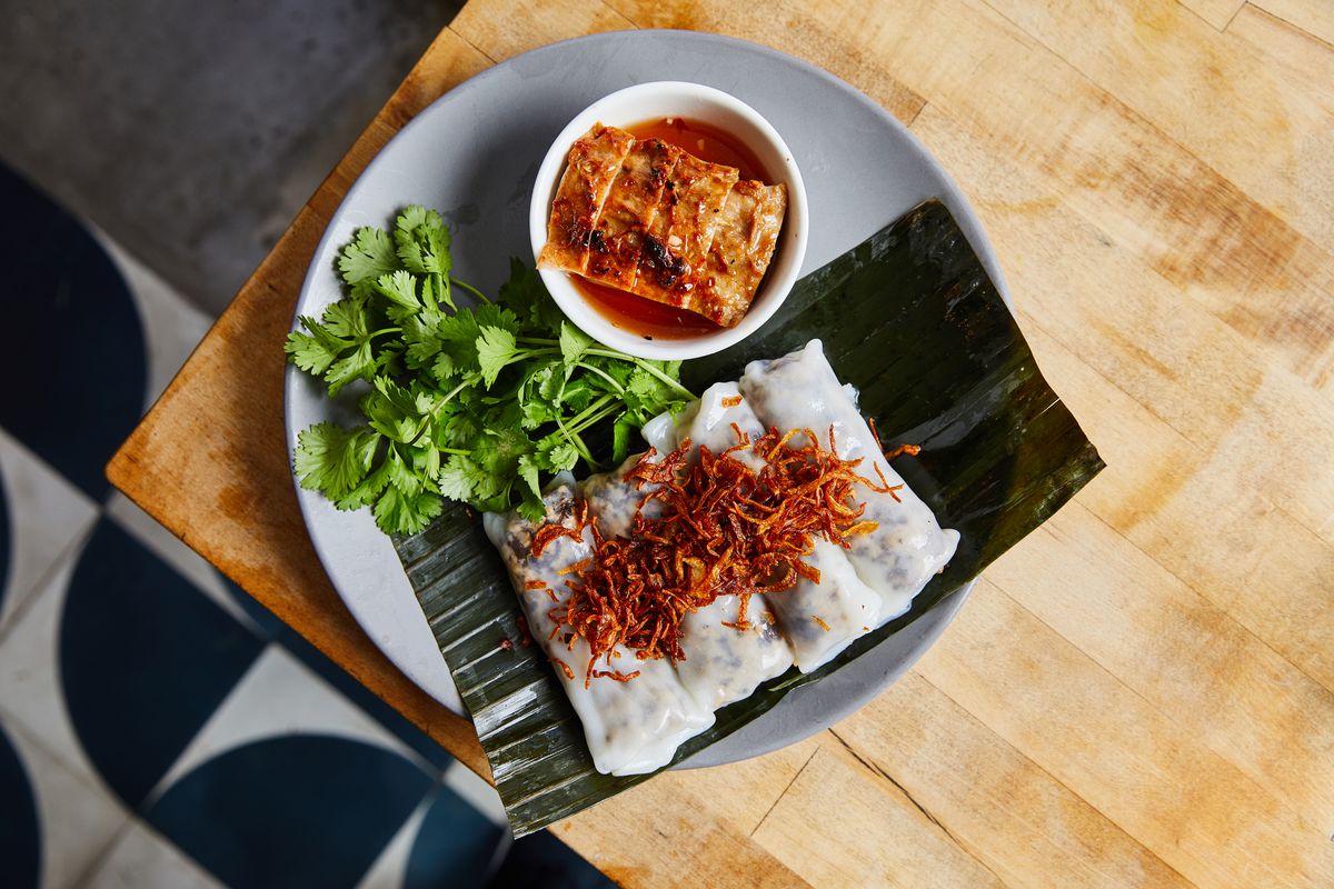 A light blue plate with cilantro, rice rolls, fried scallions, a white cup placed on top. A banana leaf is placed over part of the dish and under the food for decor. The plate is set on the edge of a light wooden table.
