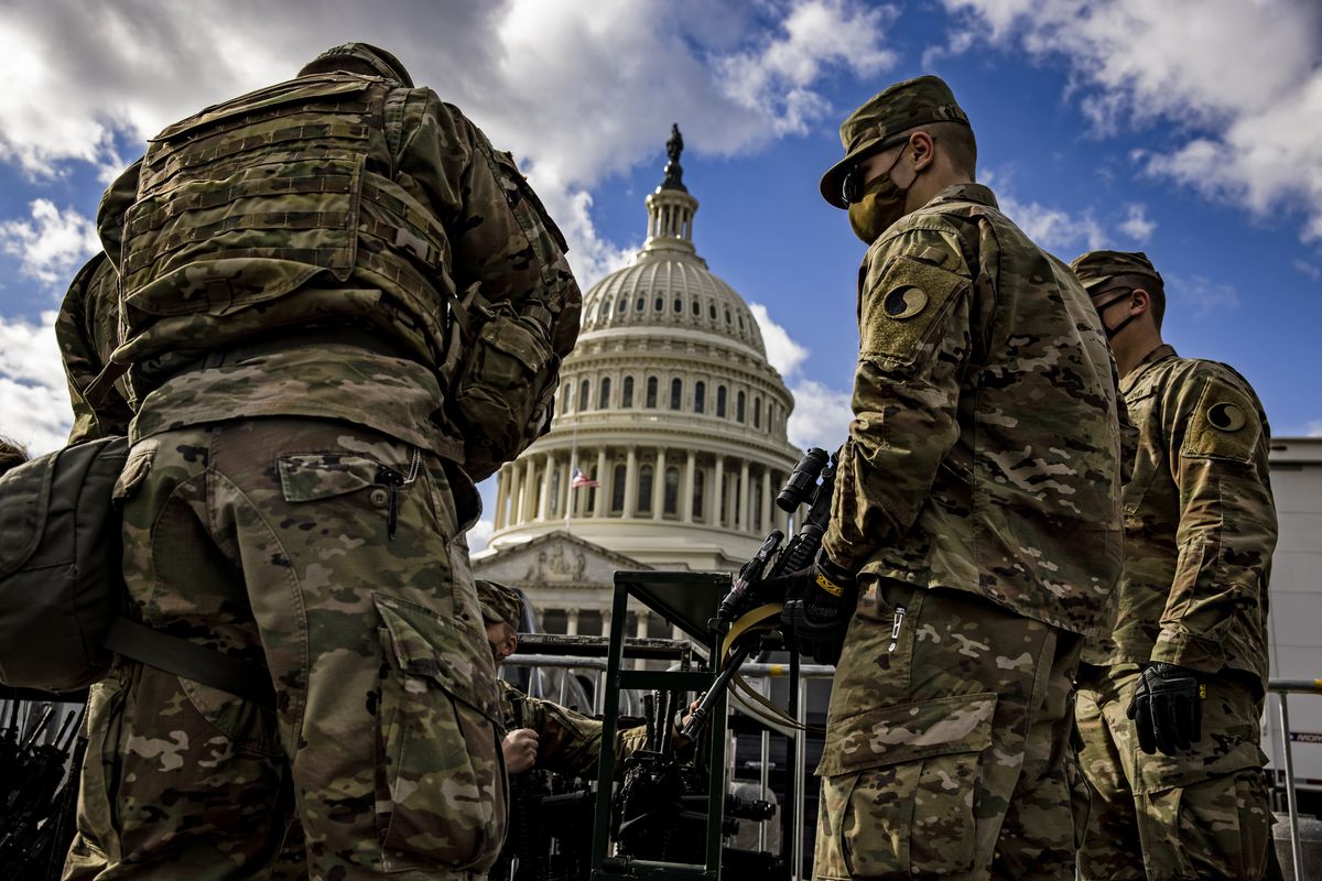 Men in green fatigues check in with a burly man in bulletproof military vest, and are being given assault rifles. Behind them looms the white dome of the Capitol building, rising up behind tall black fences.