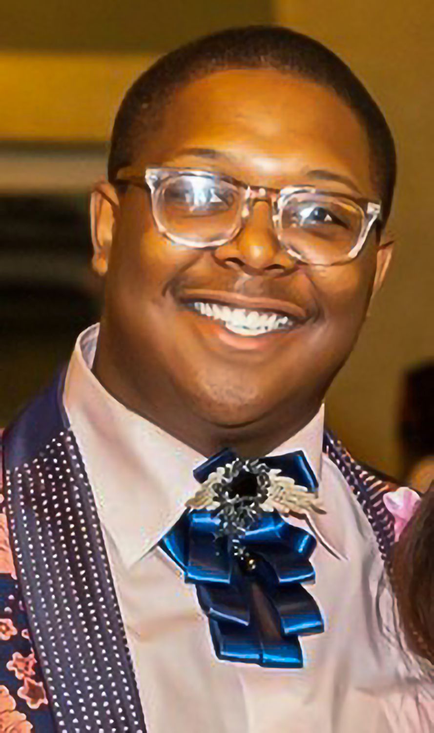 Trey Cunningham, wearing a blue frilled tie and glasses, smiles at the viewer.