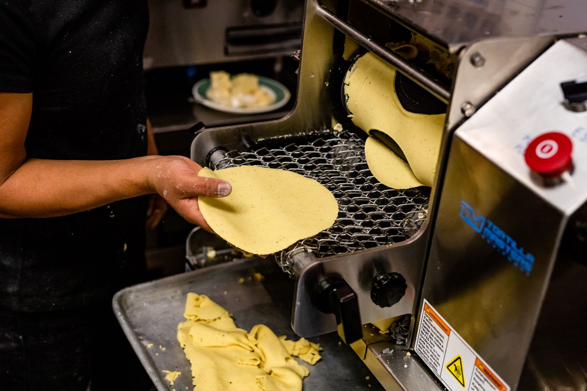 A person puts a catches a freshly made corn tortilla as it’s delivered from a machine.