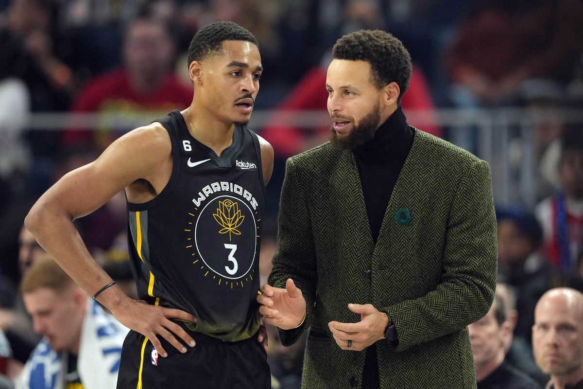 Jordan Poole, in a Warriors jersey, talking with Steph Curry, in a suit
