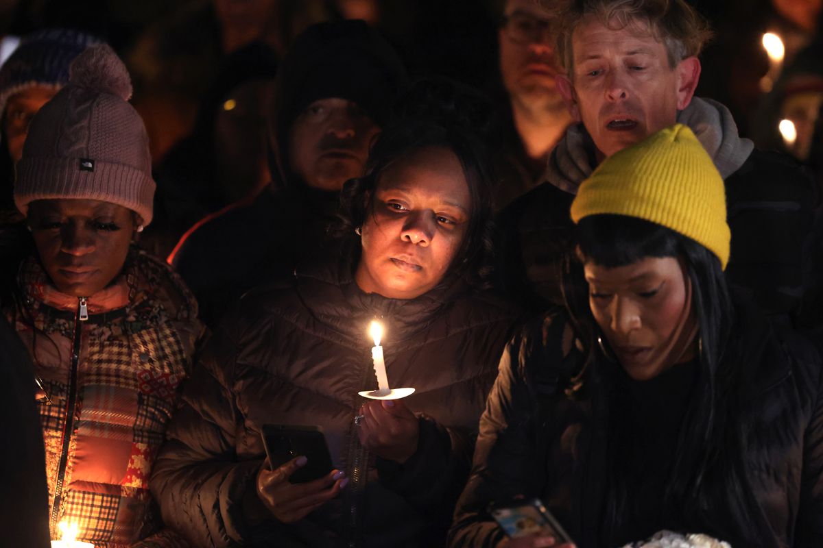 A huddled group of mostly Black faces, some partially hidden by hats and scarves, are lit by candlelight. The Black woman at the center of the photo holds a candle near her face, which seems drawn with grief. The expressions on the faces surrounding her, seen dimly though the gloom, match hers.