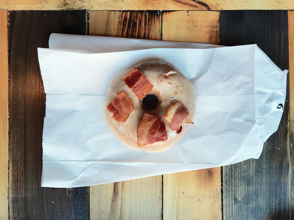 Overhead shot of a maple glazed doughnut with pieces of bacon on it. It’s sitting on a white paper bag on a rustic wooden table.