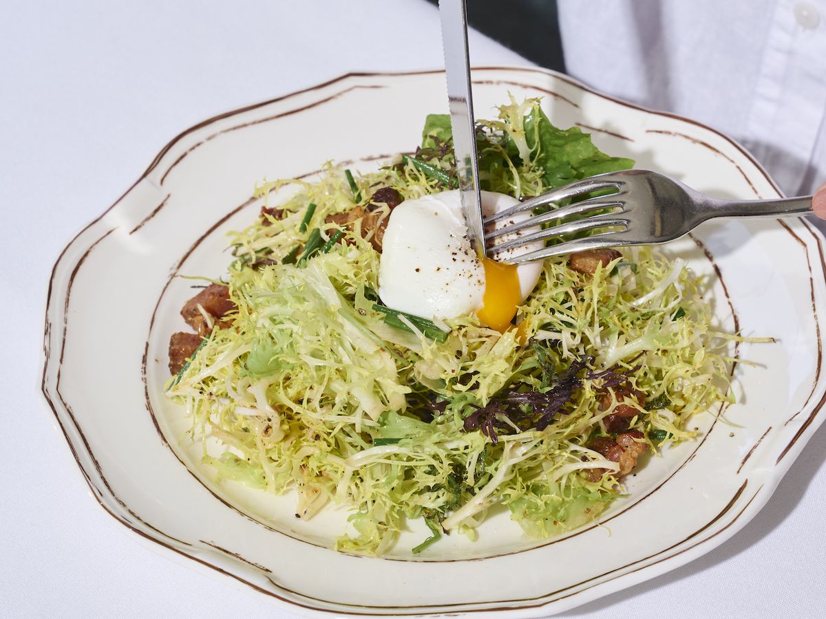 A frisee salad with soft egg with a knife going through it as a person in a white shirt cuts, inside a new restaurant Shirley Brasserie in Hollywood.