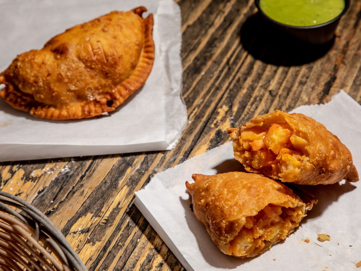 A golden Viagra empanada sits on wax paper, sliced in half, on the lower right-hand side of the photo, while a whole empanada lies on the upper left; a ramekin of green salsa sits in between