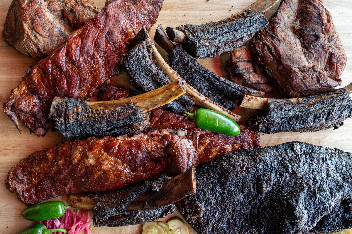 Pieces of brisket with dark bark and individual ribs are placed on a piece of butcher block, with jalapenos scattered in.