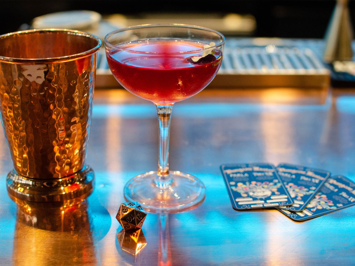 A reddish purple cocktail sits on a copper bar next to a shiny copper cup, 20-sided die, and three black cards with colorful graphics on them.
