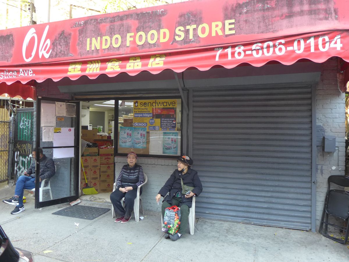 A store with a battered red awning and three people sitting in chairs in front.