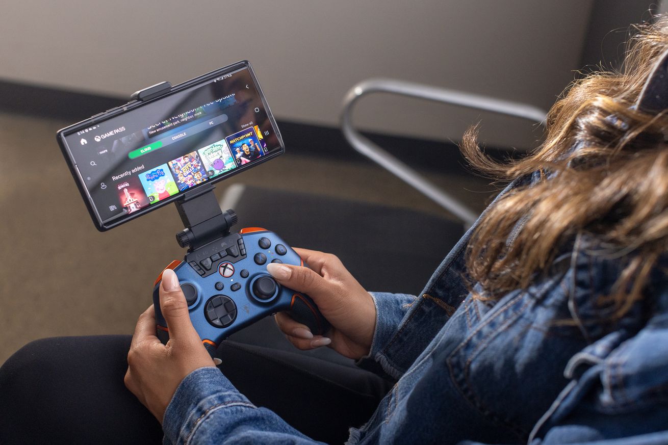 The blue Turtle Beach Recon Cloud controller with a smartphone attached to it via its included mobile phone clip. The controller and phone are in the hands of a seated person, who is using the setup to play a cloud-streamed game on Xbox Game Pass.