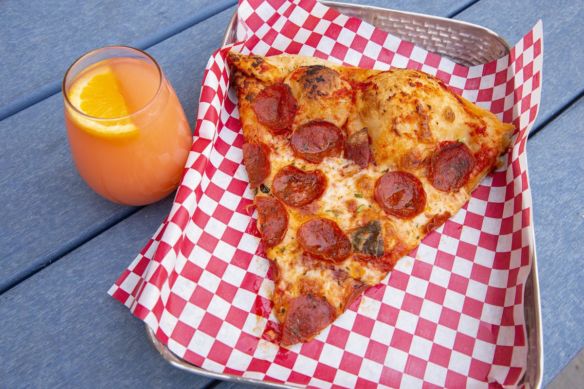 A slice of pepperoni pizza on a checkered piece of paper inside a metal basket on a picnic table beside an orange cocktail.