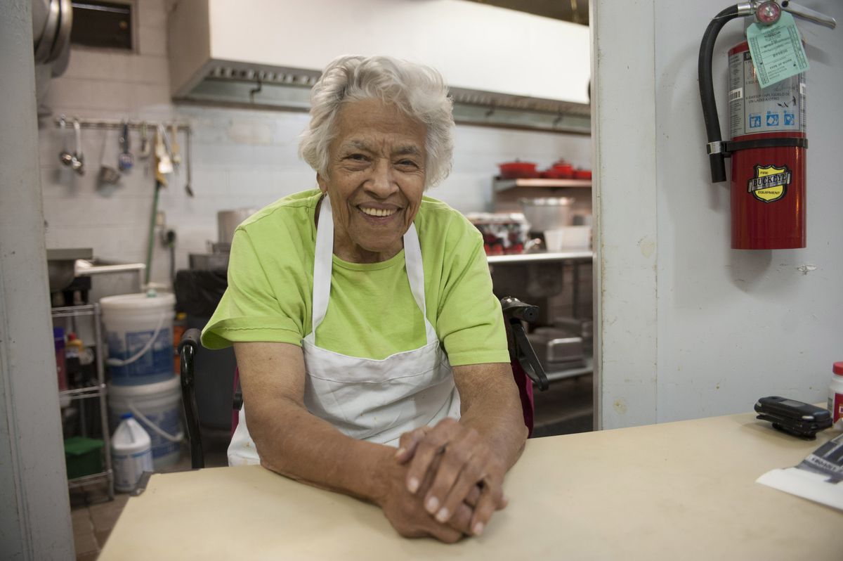 An older woman sits in a kitchen in a bright green top. Her arms rest on a butcher block.